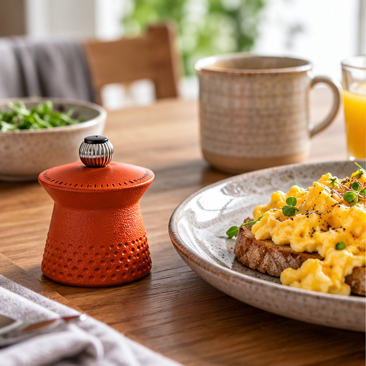 Orange Peugeot Bali Fonte cast-iron pepper mill beside scrambled eggs on toast on a sunny breakfast table